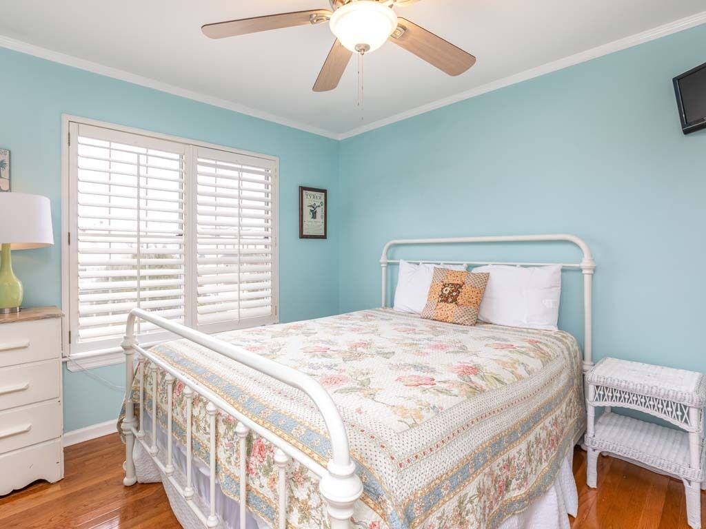 A bedroom with a bed, a ceiling fan, a window with blinds, a bedside table, and a small white chair.
