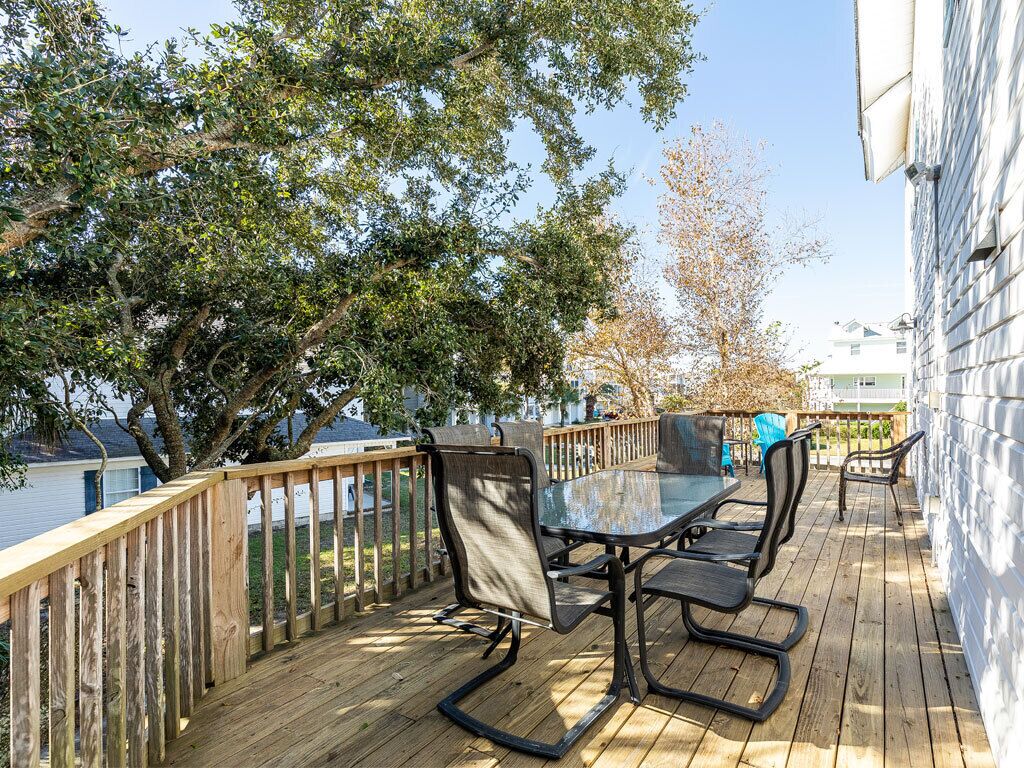 A deck with a glass-top table and chairs, surrounded by a wooden railing, with trees and a building in the background.