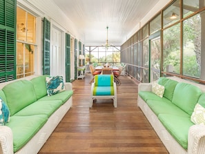 A spacious room with a wooden floor, green upholstered sofas, a dining table with chairs, and large windows offering a view of greenery.