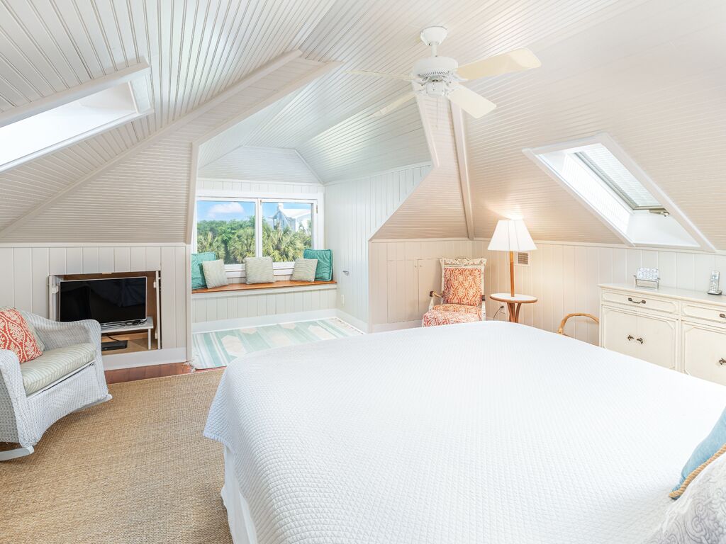 A bedroom with a large bed, a television, a chair, and a desk. There is a window with a view of greenery outside.