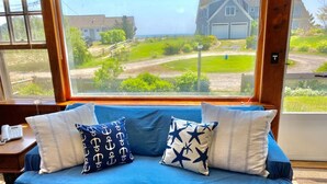A blue sofa with patterned cushions in a room with a large window overlooking a residential area. - Cozy Oceanview 1BR Cottage Across from the Beach! (Harwich)