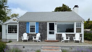 A single-story house with a grey shingle roof, a front porch with white chairs, and a small garden with purple flowers. - Cozy Oceanview 1BR Cottage Across from the Beach! (Harwich)