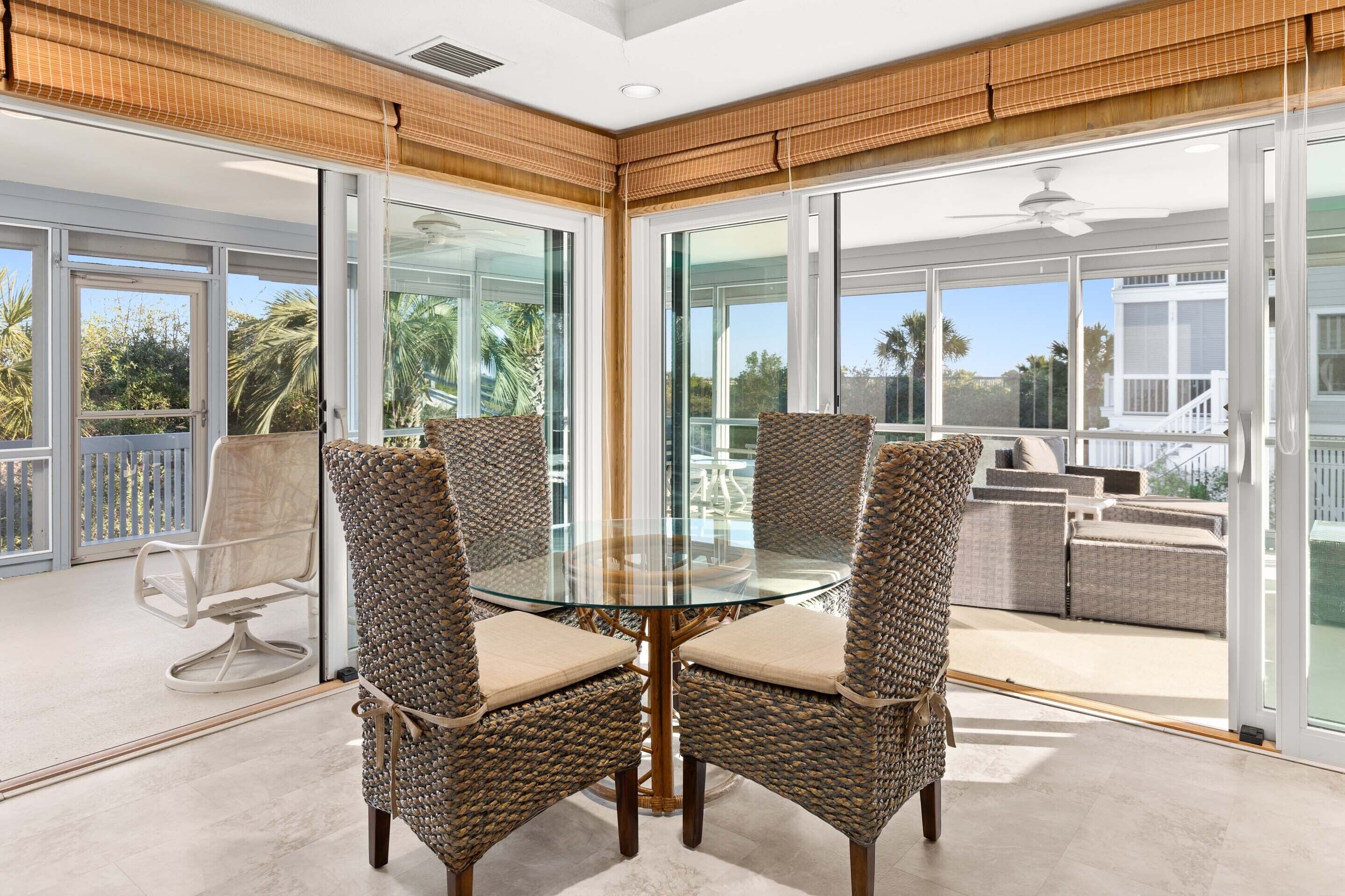 Un comedor con paredes de vidrio, sillas de mimbre y una mesa de vidrio, con vista a un balcón con mobiliario exterior.
