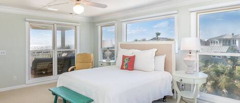 A bedroom with a large bed, a ceiling fan, a bench, a small table, and a view of the outdoors through large windows.