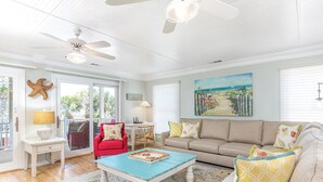 A spacious living room with a red sofa, beige sofa, and a blue coffee table. There is a ceiling fan, a painting on the wall, and a starfish decoration.
