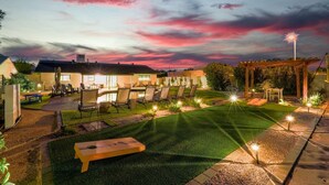 A well-lit outdoor patio area with a lawn, seating arrangements, and a pergola. - Upscale Oasis | Pool, Putting Green, Volley Court & Open Yard (Scottsdale)
