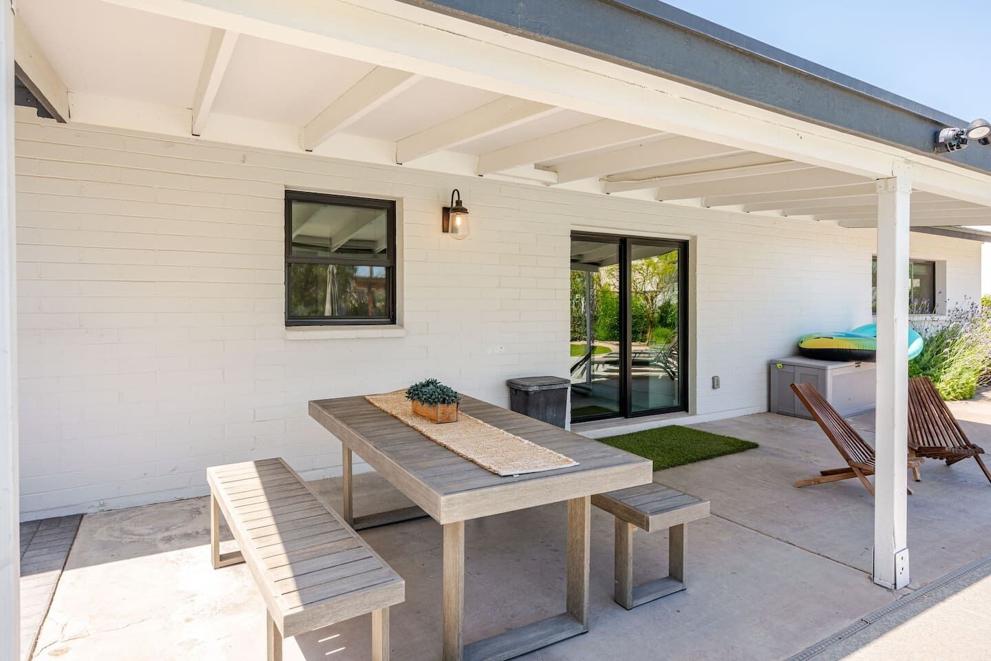 A covered patio with a wooden table and benches, a small potted plant, and a grill area.