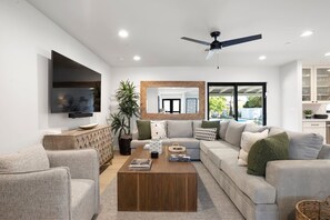 A modern living room with a flat-screen TV, a ceiling fan, a large mirror, and a view to the outside. - Upscale Oasis | Pool, Putting Green, Volley Court & Open Yard (Scottsdale)