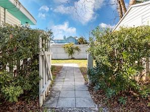 A narrow concrete pathway flanked by dense green foliage, leading to a residential area with a white fence and a blue house in the background.