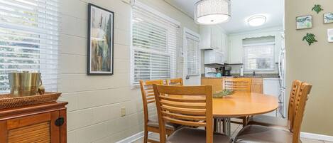 A dining area with a round table and wooden chairs, a kitchen in the background, and a window with blinds.
