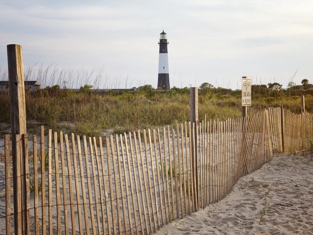 Une clôture en bois avec un panneau, une plage de sable et un phare en arrière-plan.