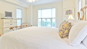 Une chambre spacieuse avec un grand lit, un ventilateur de plafond, un téléviseur posé sur une commode et une fenêtre équipée de stores.