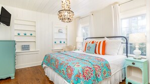 A bedroom with a bed, a chandelier, a nightstand, and a window. - Sassy Seagull by Tybee Vacation Rentals (Tybee Island)