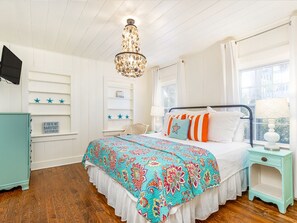 A bedroom with a bed, a chandelier, a nightstand, and a window. - Sassy Seagull by Tybee Vacation Rentals (Tybee Island)