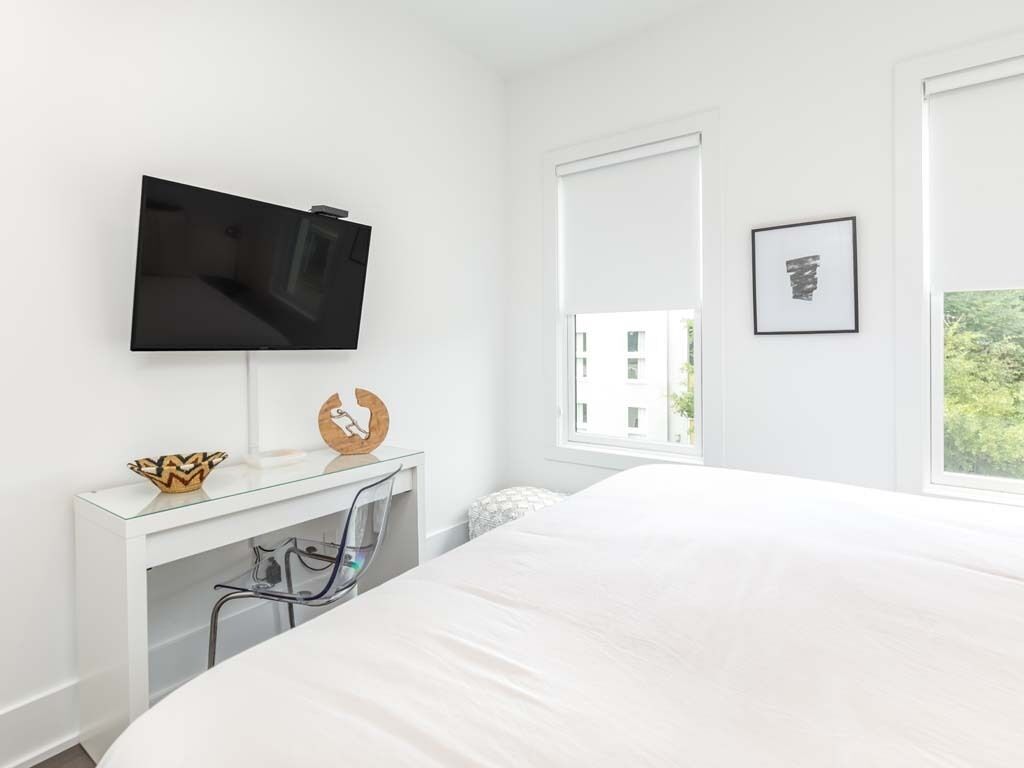 Ein modernes Schlafzimmer mit Bett, einem an der Wand befestigten Fernseher, Schreibtisch mit Stuhl und einem Fenster mit Jalousien.
