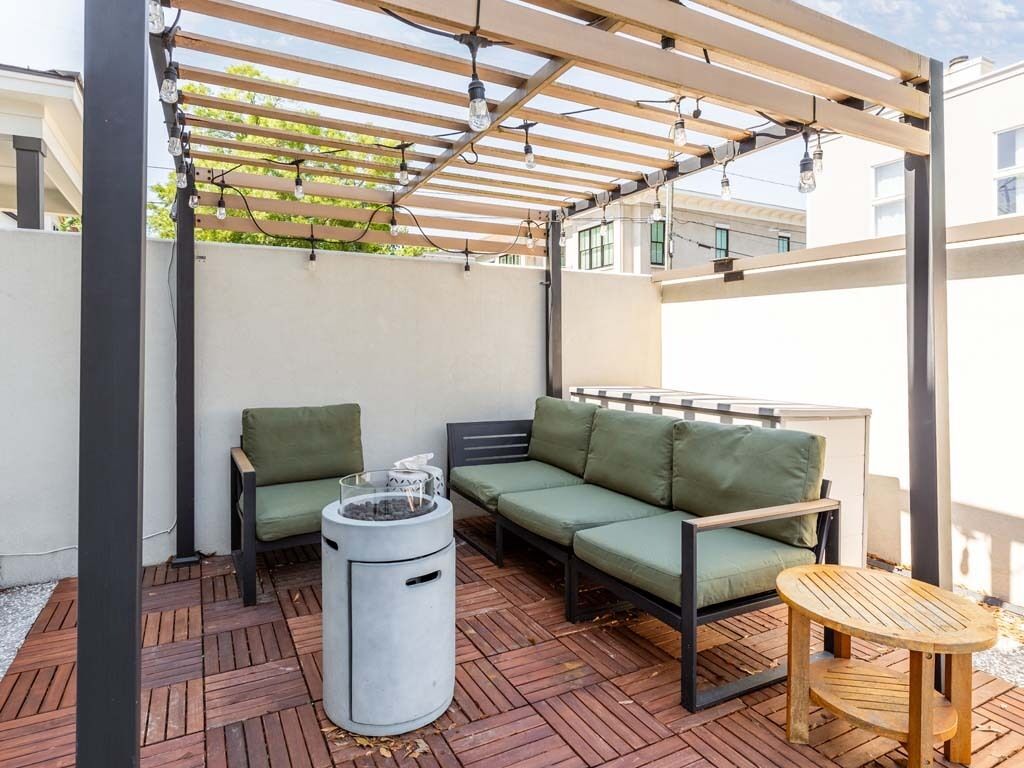 An outdoor patio with a wooden pergola, a green sofa, a round table, and a trash bin.