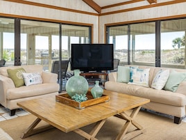 A living room with two sofas, a coffee table, a TV, and large windows offering a view of the landscape.