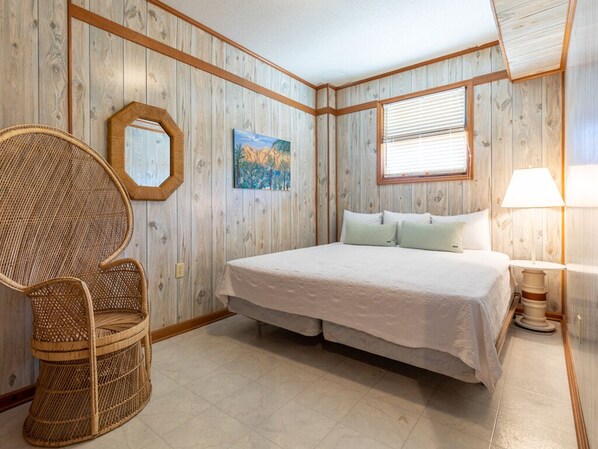 A bedroom with a large bed, a wicker chair, a painting, and a window with blinds. - Sea Gull Ocean Villa by Tybee Vacation Rentals (Tybee Island)