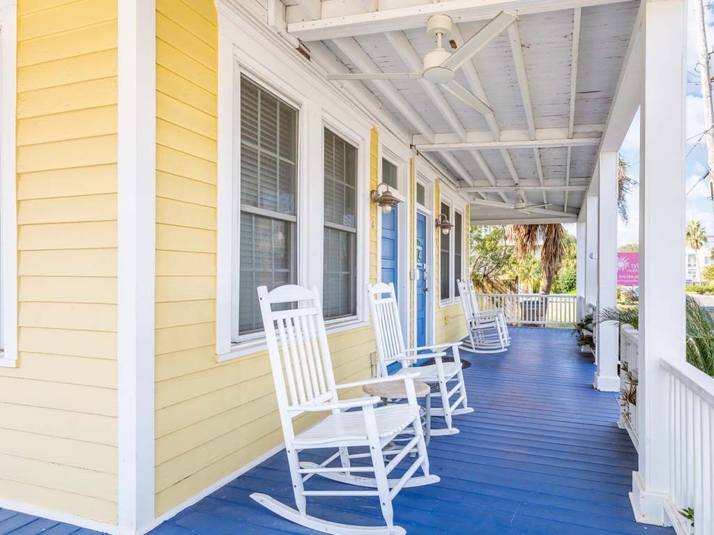 A porch with white rocking chairs, a blue floor, and a yellow building.