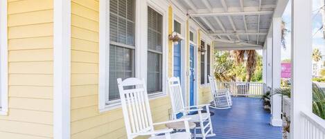 A porch with white rocking chairs, a blue floor, and a yellow building.
