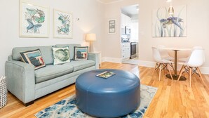 Un salon comprenant un canapé, un pouf et un coin repas avec des chaises. Les murs sont ornés d’œuvres d’art encadrées et le plafond est équipé d’un ventilateur.