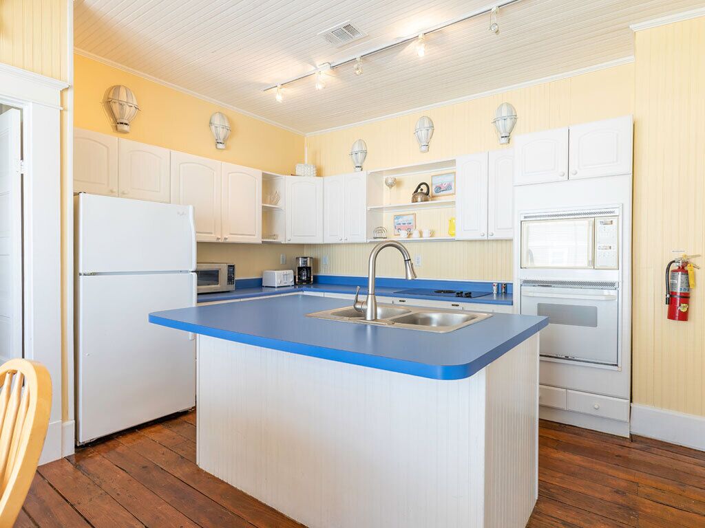 A compact kitchen with white cabinets, a blue countertop island, a stainless steel sink, a microwave, a stove, and a fire extinguisher.