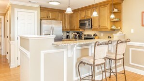 A kitchen with wooden cabinets, a stainless steel refrigerator, a microwave, and a bar area with two bar stools. - Silver Seas 203 by Tybee Vacation Rentals (Tybee Island)