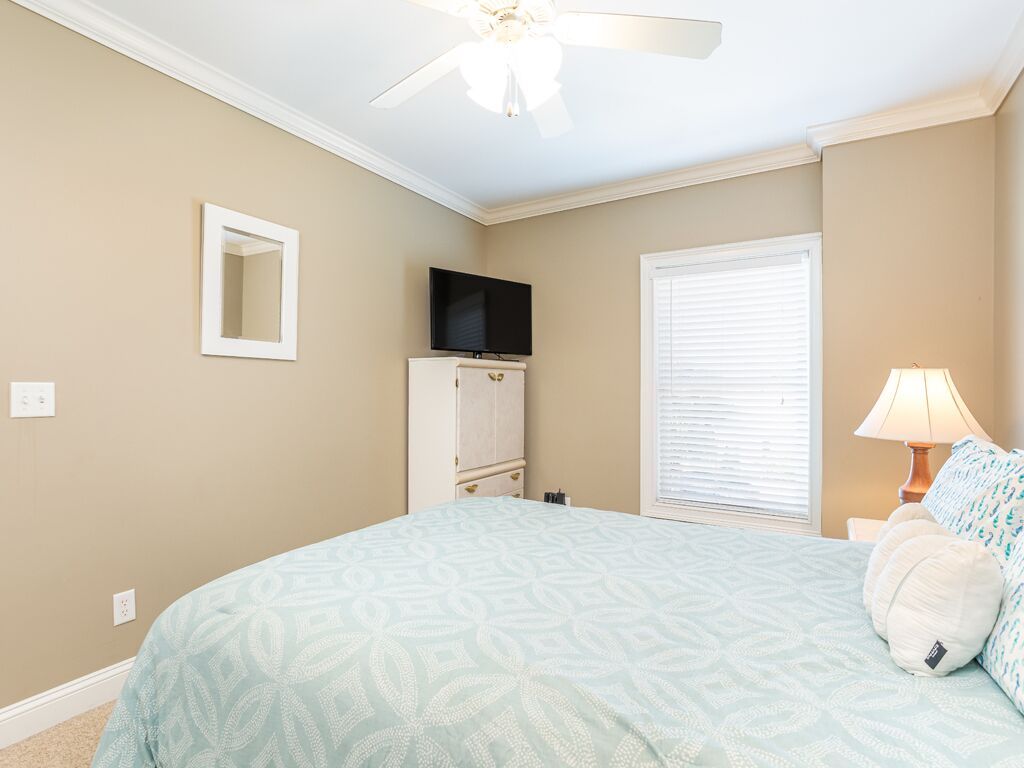A bedroom with a bed, a ceiling fan, a television, a window with blinds, and a nightstand with a lamp.