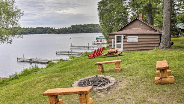 Una cabaña junto a un lago, con un brasero y mesas de picnic.