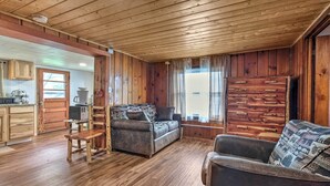 Interior de una cabaña de madera con sofá, sillón, un mueble de madera y una zona de cocina.