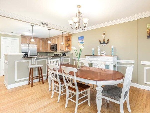 A dining area with a wooden table and white chairs, a kitchen with wooden cabinets and stainless steel appliances, and a chandelier.