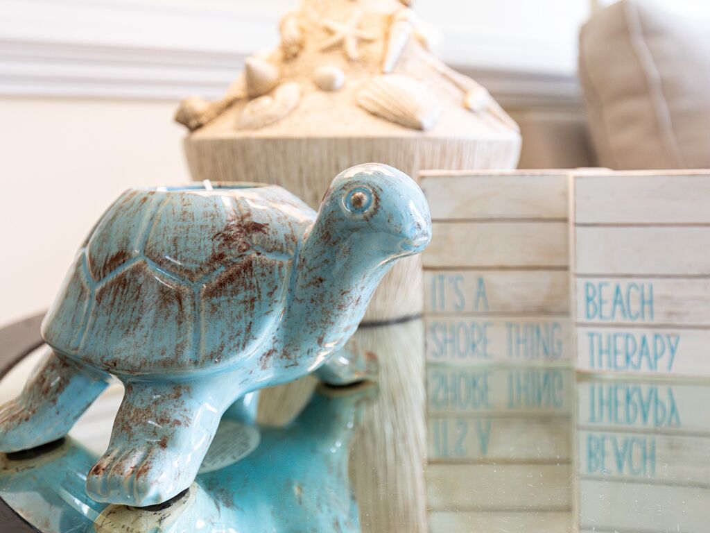 Une figurine de tortue en céramique est posée sur une table. En arrière-plan, on aperçoit une boîte en bois avec du texte et un objet décoratif orné de coquillages.