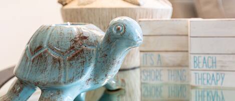 Une figurine de tortue en céramique est posée sur une table. En arrière-plan, on aperçoit une boîte en bois avec du texte et un objet décoratif orné de coquillages.