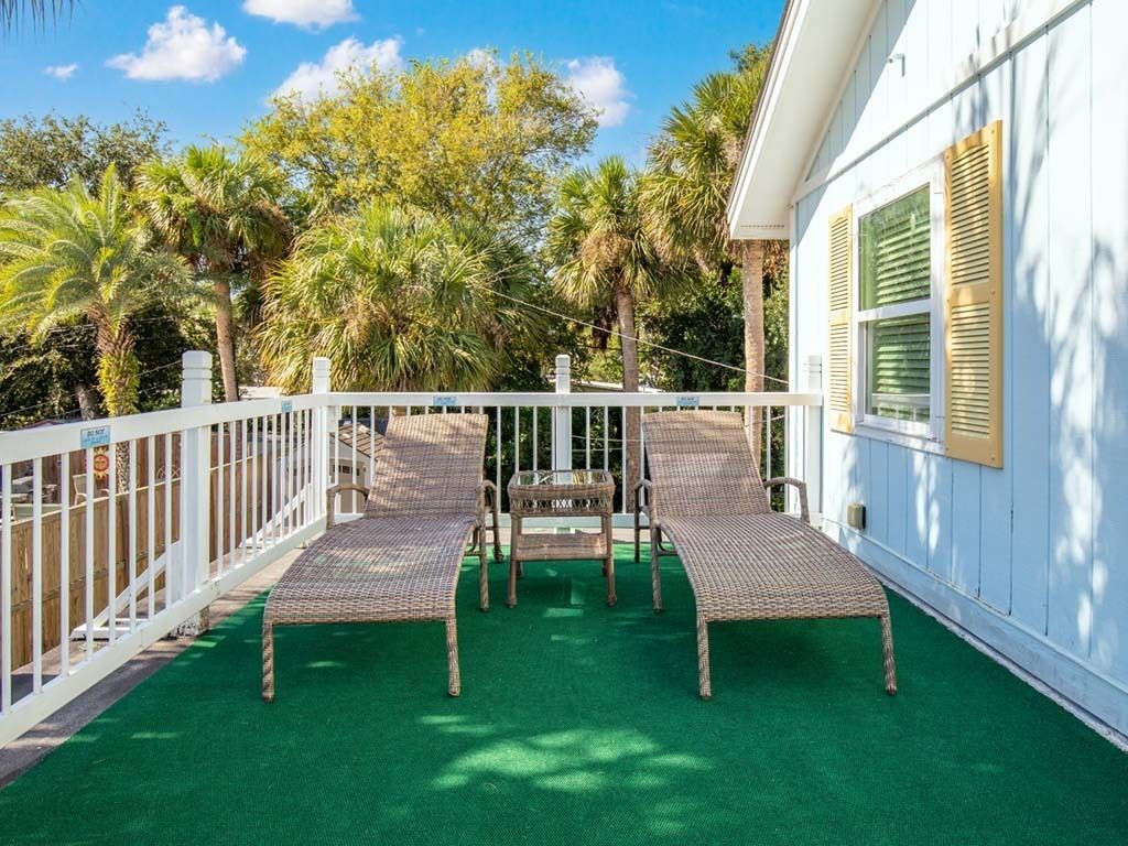 A balcony with two wicker lounge chairs and a small table, surrounded by a white fence and palm trees.