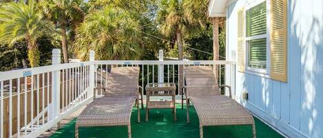 A balcony with two wicker lounge chairs and a small table, surrounded by a white fence and palm trees.