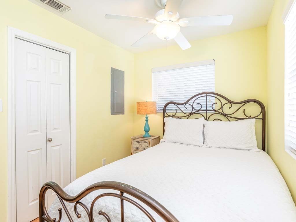 A bedroom with a bed, a ceiling fan, a window with blinds, a nightstand with a lamp, and a door.