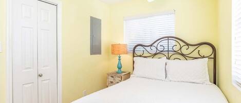 A bedroom with a bed, a ceiling fan, a window with blinds, a nightstand with a lamp, and a door.