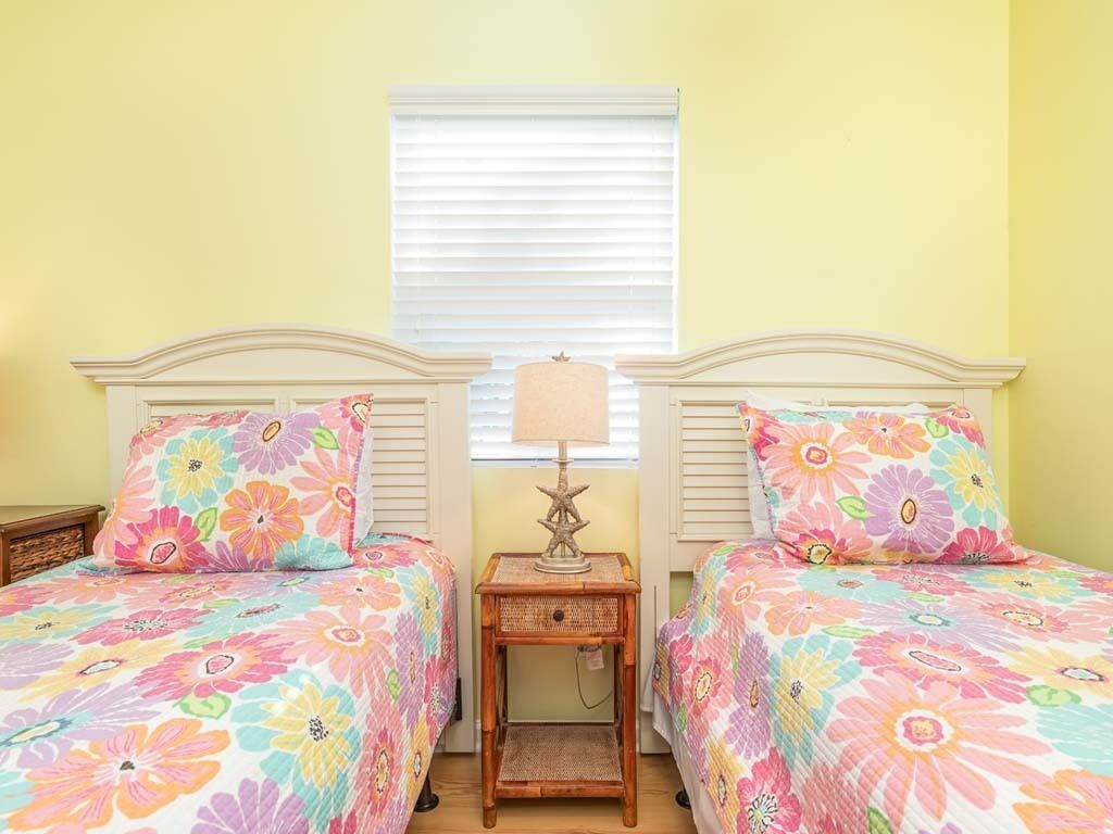 A room with two beds, each with floral bedding, a small wooden nightstand with a lamp, and a window with blinds.