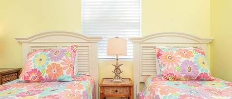 A room with two beds, each with floral bedding, a small wooden nightstand with a lamp, and a window with blinds.