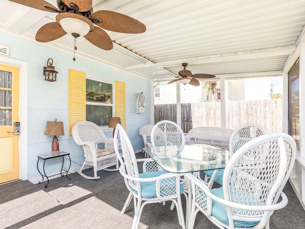 Une terrasse couverte avec une table ronde en verre et des chaises en osier blanches, un ventilateur de plafond et une porte dotée d’un luminaire.