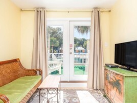 Une pièce comprenant un canapé, une petite table, un téléviseur posé sur un meuble, et une porte donnant sur un espace extérieur avec une piscine et une vue sur les arbres.