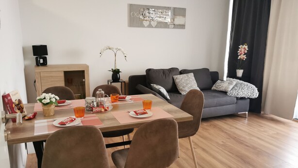 Een moderne woonkamer met een eethoek. Er is een houten eettafel met vier stoelen, een grijze bank met gekleurde kussens en een koffieapparaat op een bijzettafeltje.