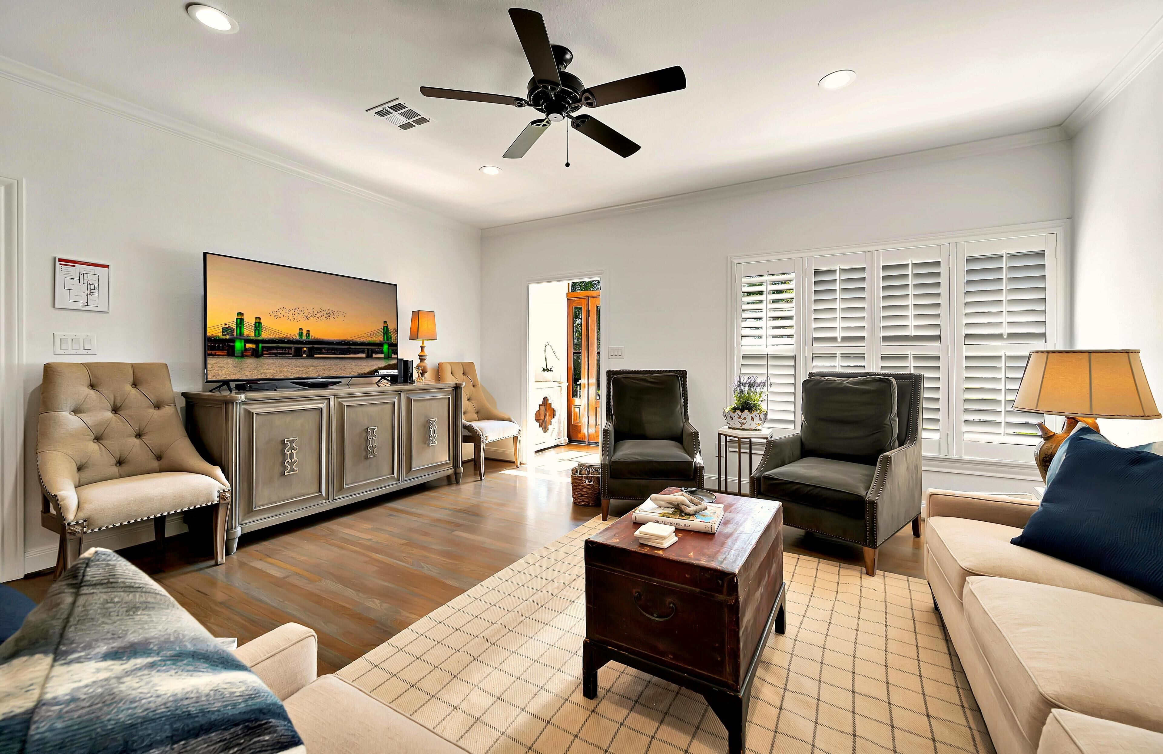 A spacious living room with a flat-screen TV, comfortable seating, a ceiling fan, and a view through the window.