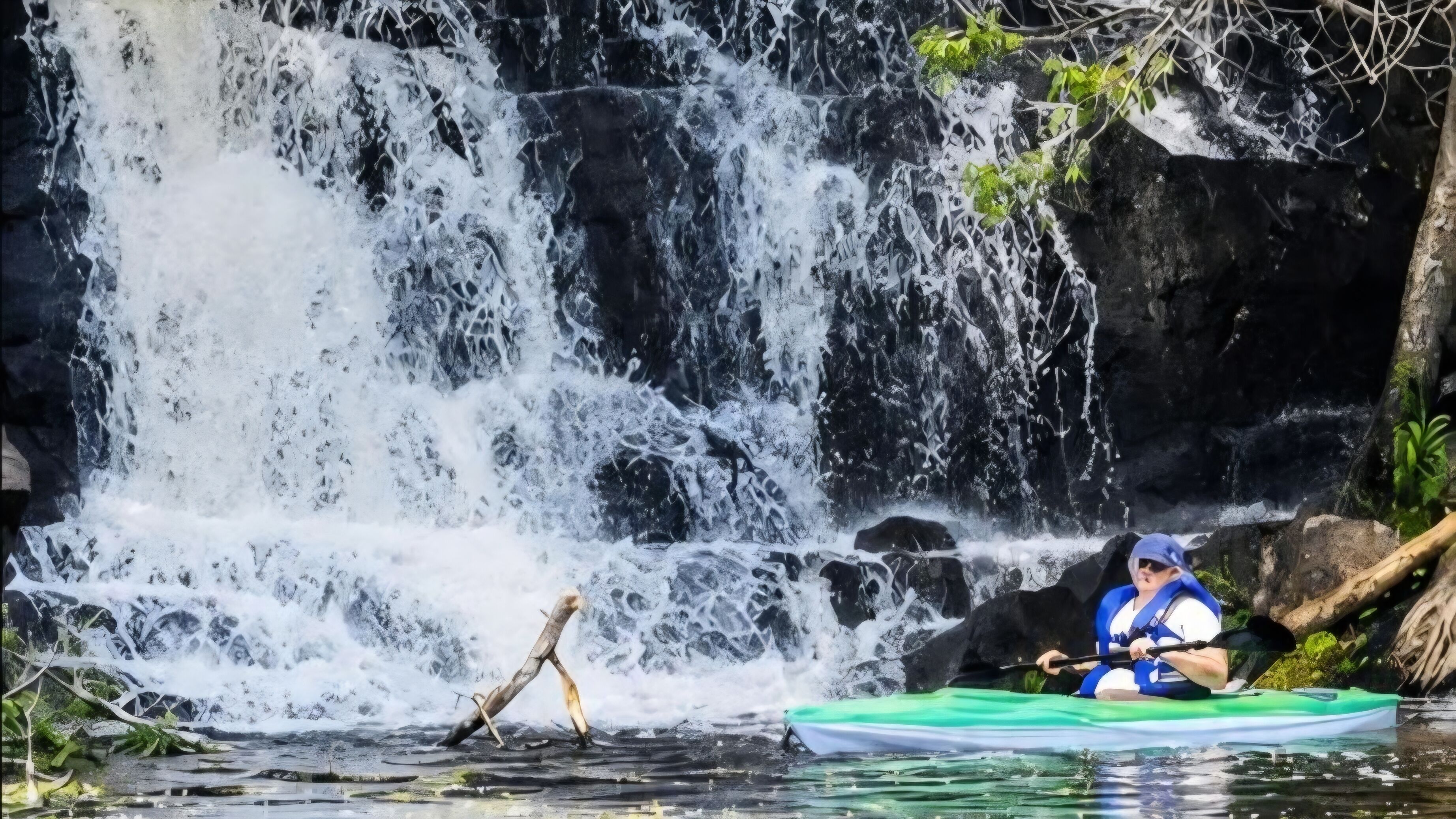 A kayaker in a green kayak on a calm river with a waterfall in the background.