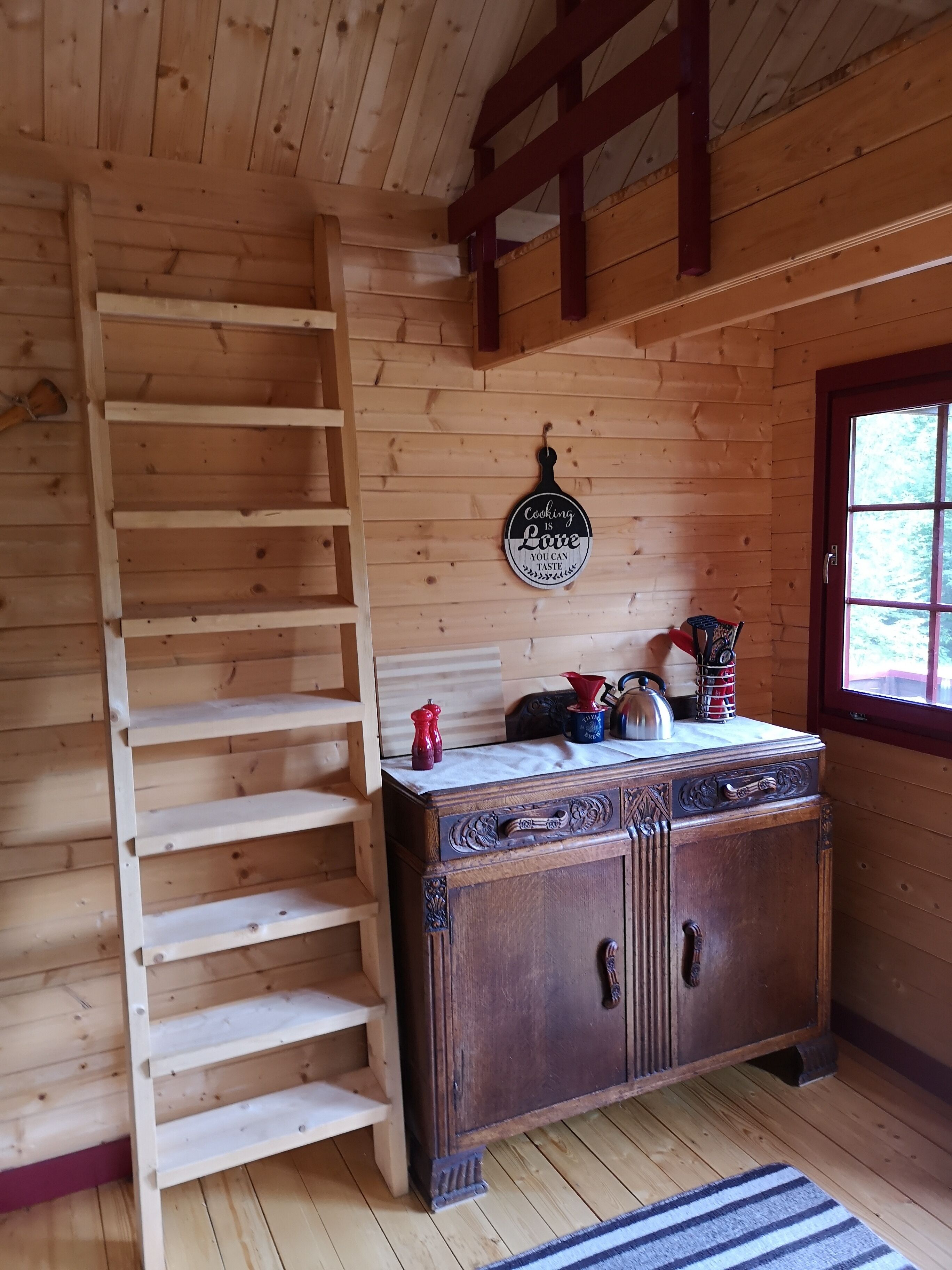 A wooden interior with a wooden ladder, a cabinet with decorative handles, and a window with a view of greenery.