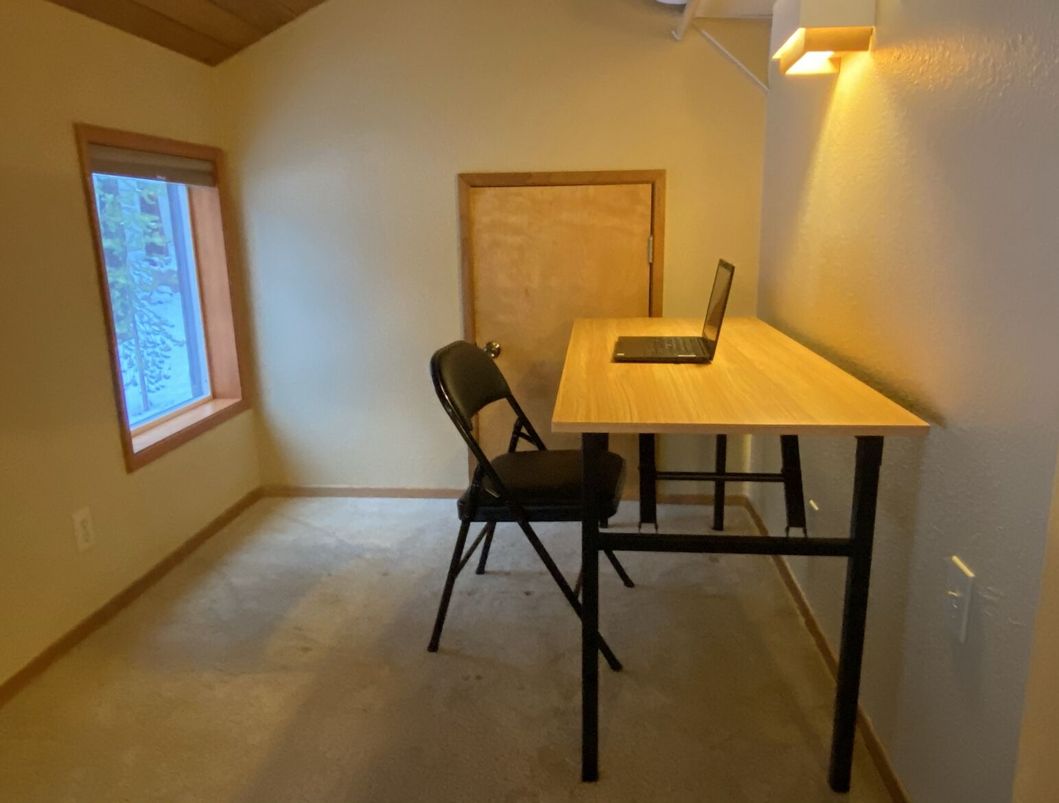 A room with a wooden desk, a laptop, a chair, and a window with a view of trees.