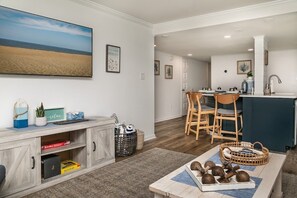 A modern living room with a large TV, a dining area with wooden chairs, and a kitchen with a blue countertop.
