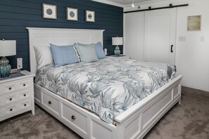 A bedroom with a white bed, blue and white floral bedspread, a white nightstand with a lamp, and a white dresser with drawers.