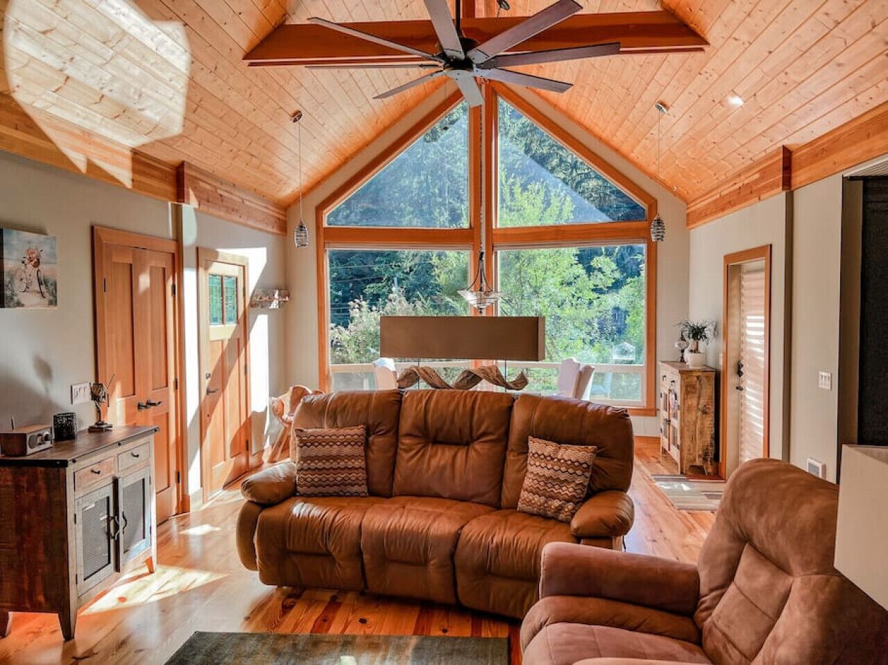 A spacious living room with a large brown leather sofa, wooden ceiling beams, and a large window offering a view of the outdoors.