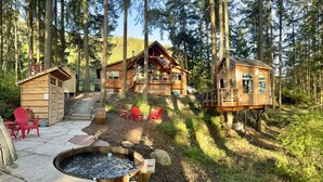 A cabin with a deck, a fire pit, and red chairs in a forest setting.